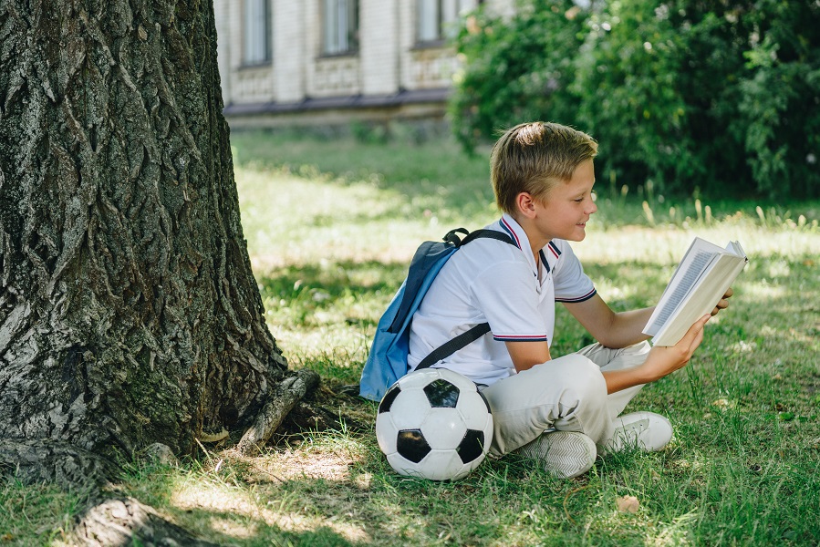 Cute,Smiling,Schoolboy,Reading,Book,While,Sitting,On,Lawn,Near